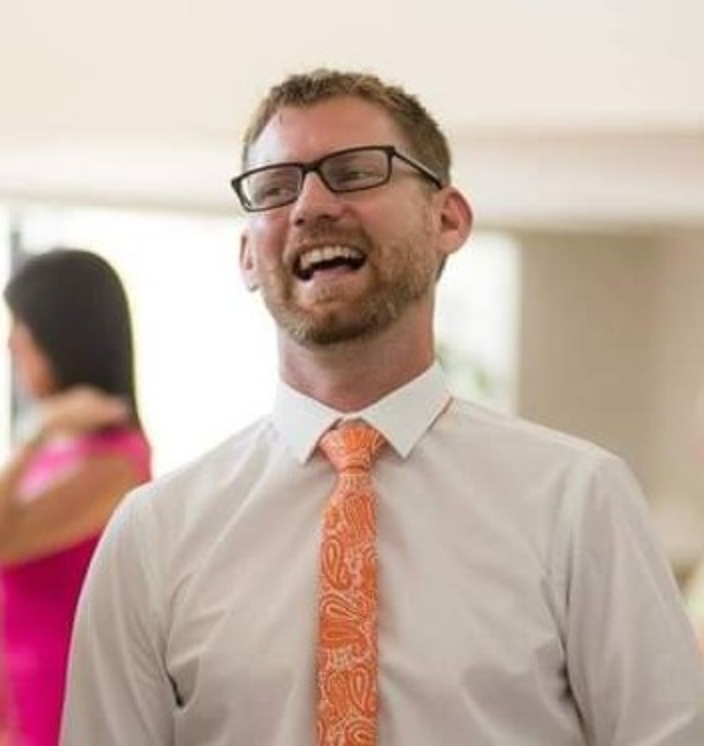 Photograph of Matthew Day smiling, wearing a white shirt and orange tie